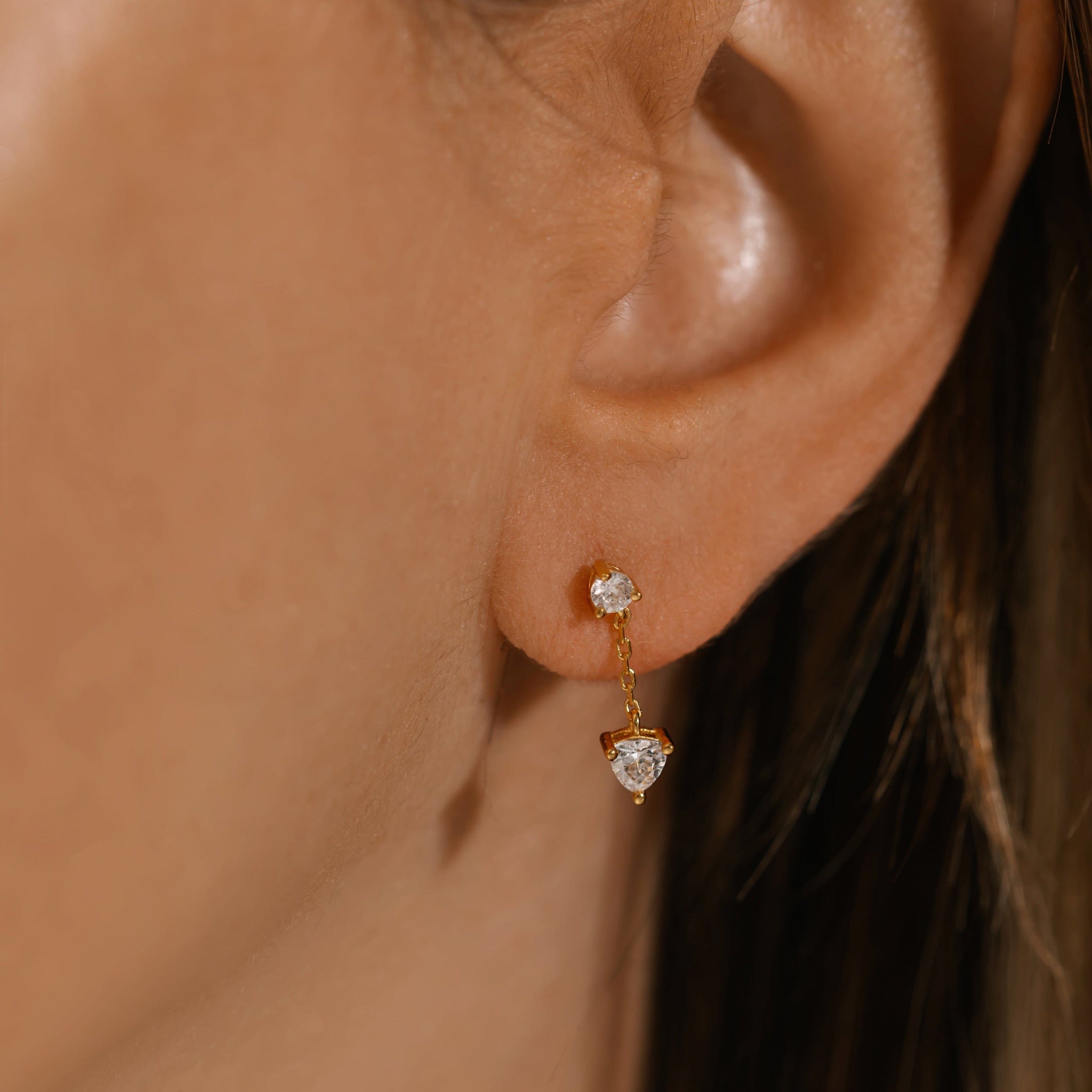 A close-up view of the Scintilla Chain Drop earring on the model's ear, the larger crystal charm dangling from the smaller crystal stud as they are connected by a thin golden chain. 
