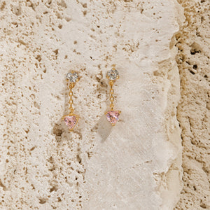 A pair of Bambina Chain Drop earrings is displayed on a block, the clear crystal accent sparkling in the light as the golden chain and pink crystal charms descended down the side of the block