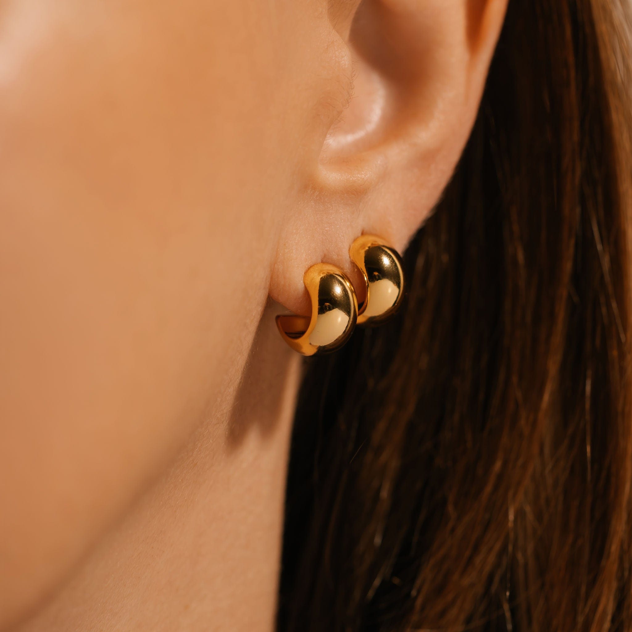 The Nice Huggie Duo is worn together on the model's ear, the golden faces of the broad, open hoops shimmering side-by-side. 