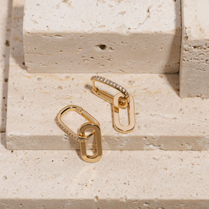 Two Positano Drop Hoop earrings lay on tiered stone slabs, the two oval hoops of each earring linked loosely together as the golden surface and crystal accents glisten brightly. 