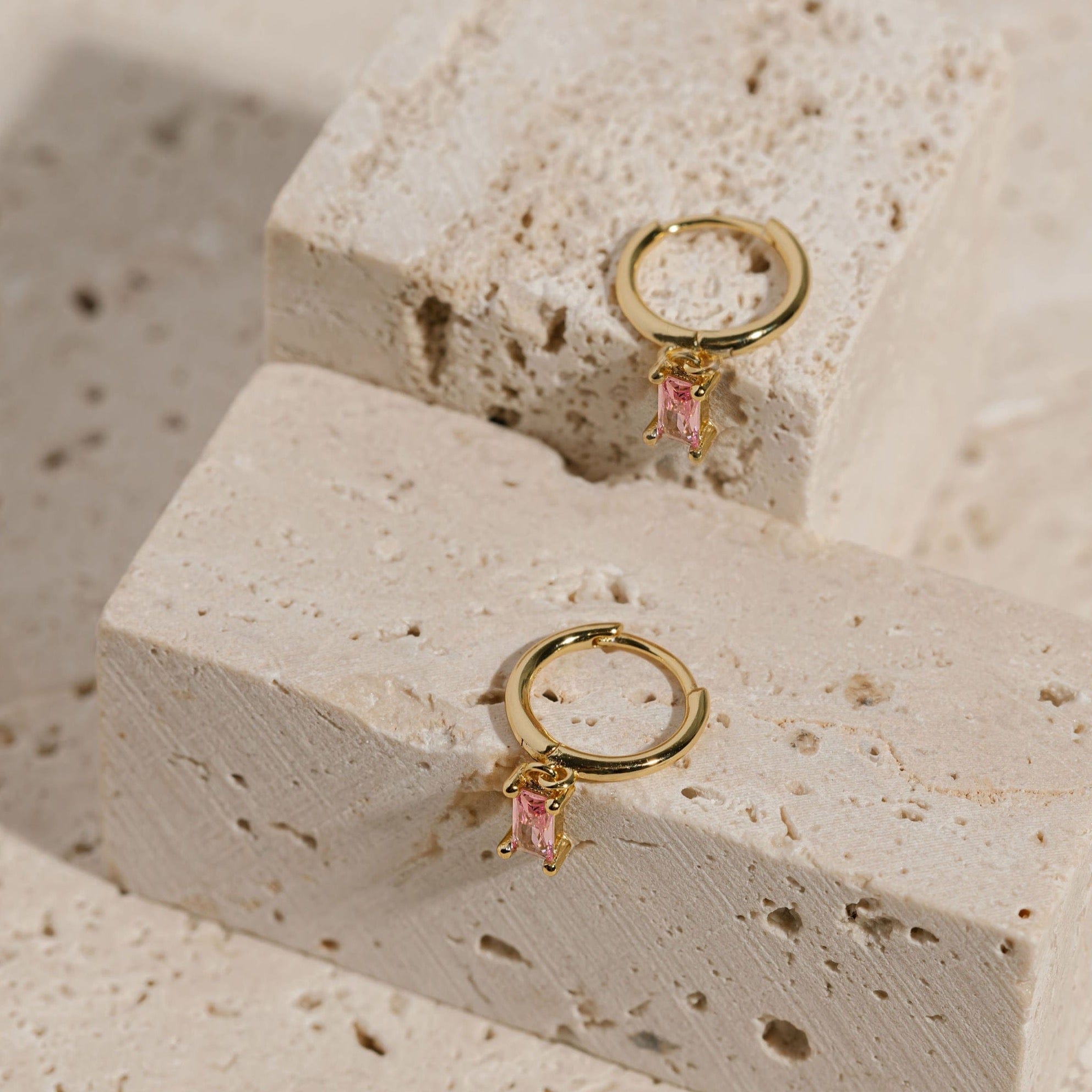 A pair of Bambina Drop Hoop earrings is displayed on two separate tiered counters, the pink crystal charm dangling down the side as the golden ring lays on top.