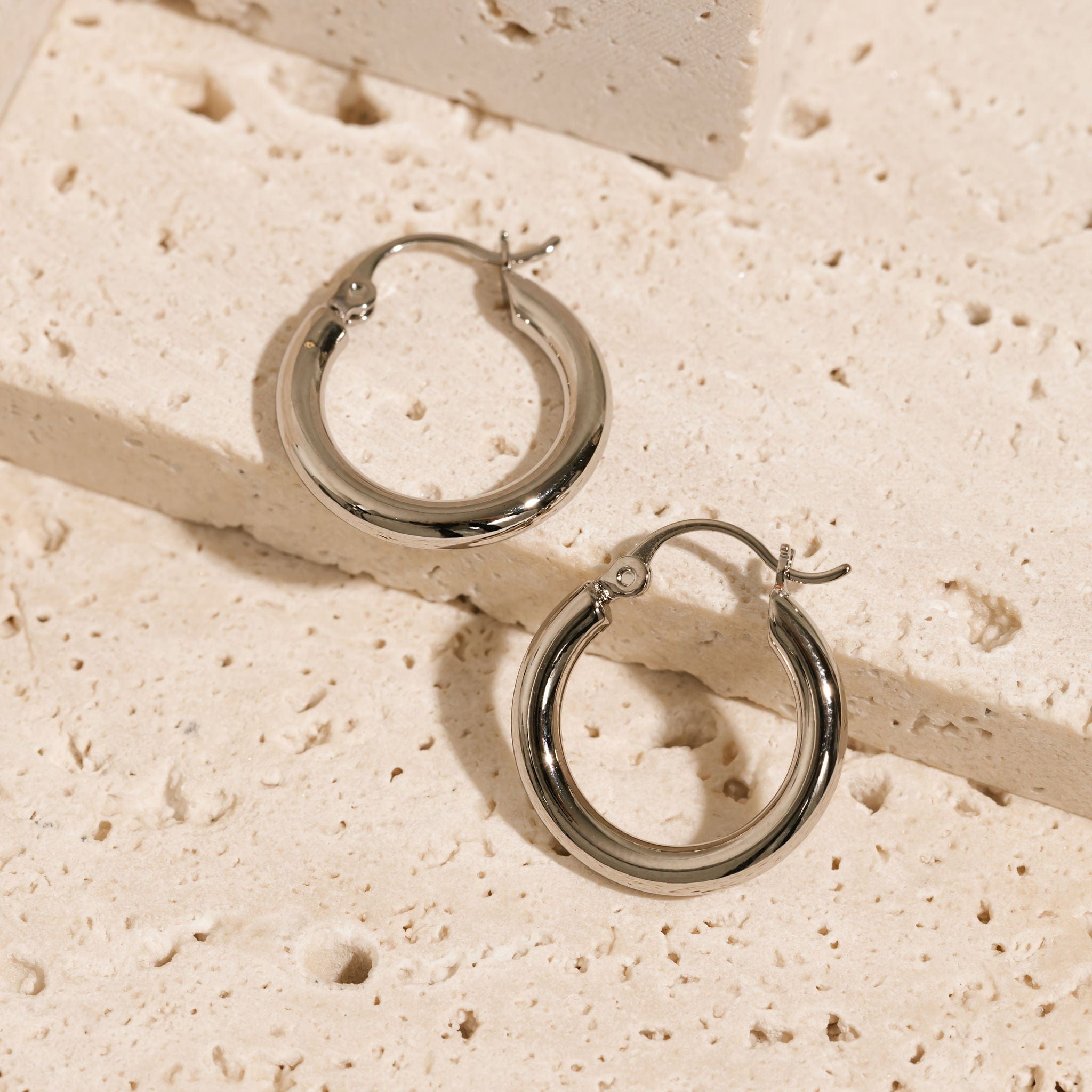 A pair of Ravello Hoop Platinum earrings lays side-by-side on a tiered stone surface to display the sleek platinum rings and latch closure. 