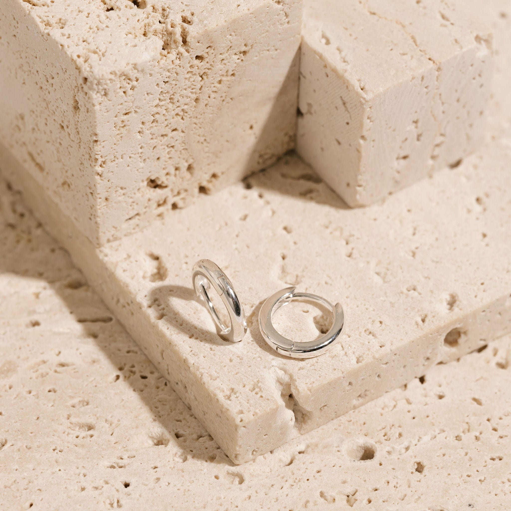 A pair of sterling silver Ravello Huggie earrings is displayed side-by-side on a tiered stone surface, one earring using the porous surface to balance on its clasp closure while the other lays flat on its side. 