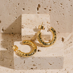 A pair of Marbella Hoop earrings is displayed on a sttone slab, the hammered texture creating light and dark reflections on the hoop. One earring lays on its side while the other is propped up to show the open C-curve of the hoop. 