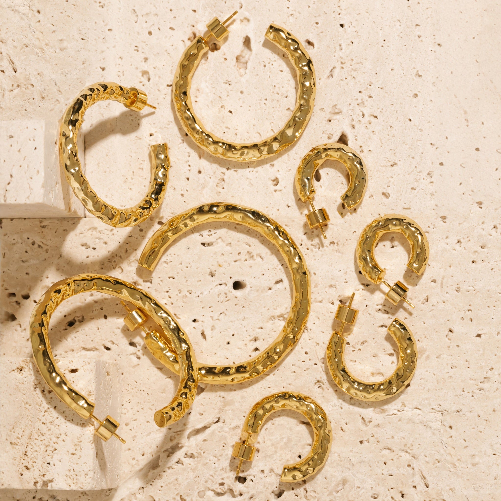 The four pairs of earrings in the Marbella Hoop Quattro set lay together on a porous stone slab to display their hammered texture and open hoop design. 