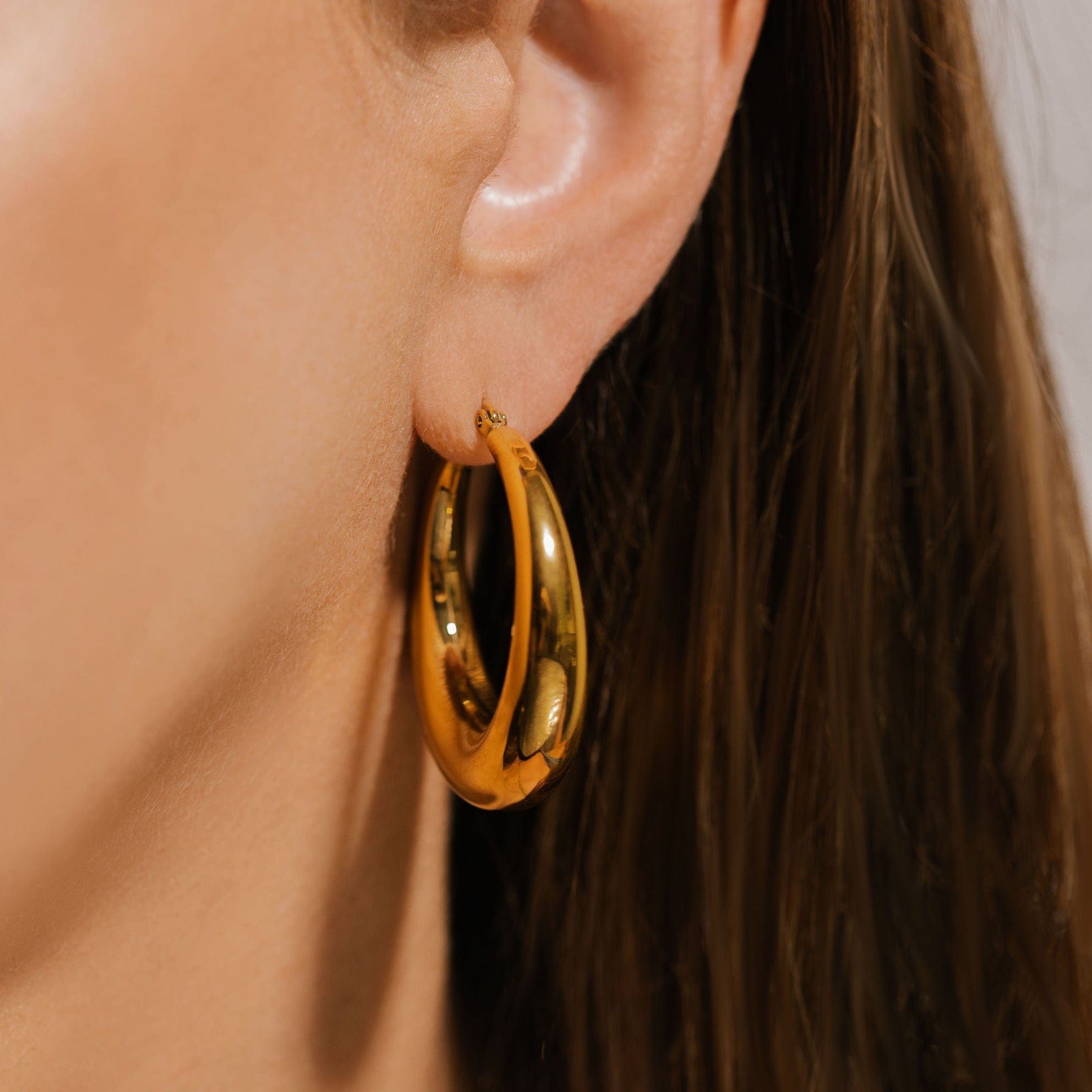 The bold face of the Capri Hoop Grande glistens and reflects the room in a close-up view of the earing on the model's ear. 
