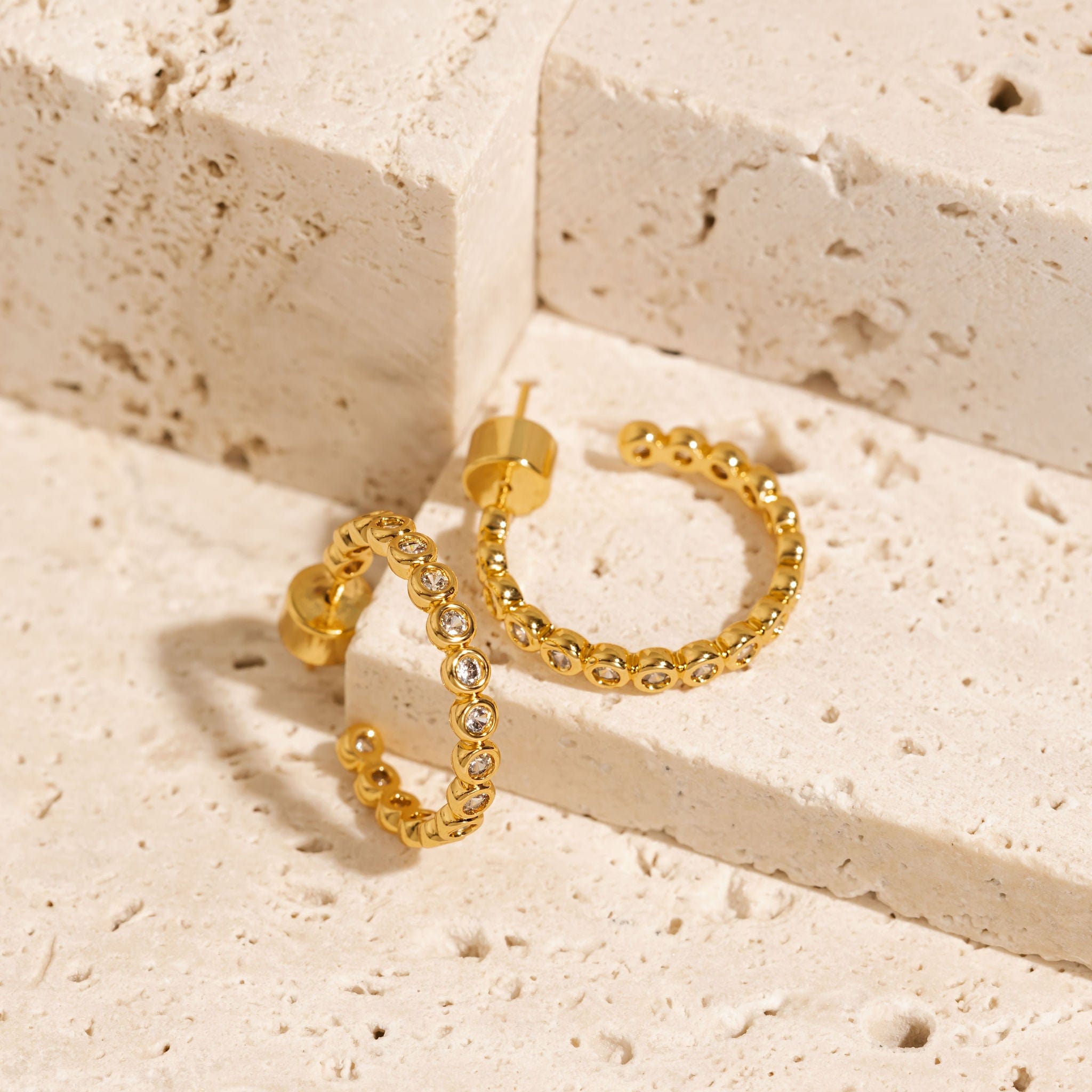 A golden Coquille Hoop earring lays on a counter to show off the gold casing and open hoop shape while another is stood up to display the sparkling crystals accents. 