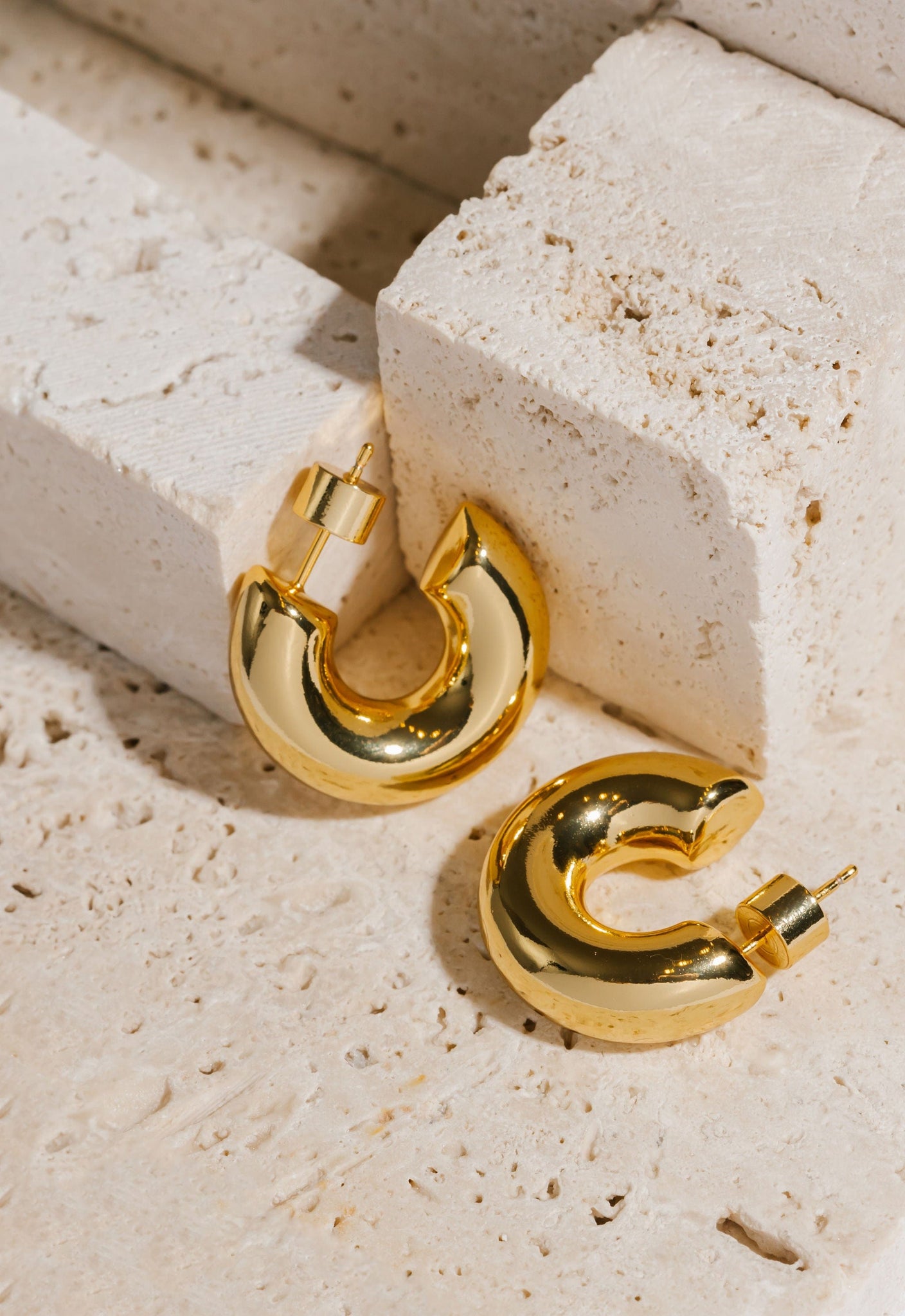 A pair of Perfect Hoop Grassetto Midi earrings lay side by side on a stone slab, their smooth and chunky open hoop design shining golden in the display light. 