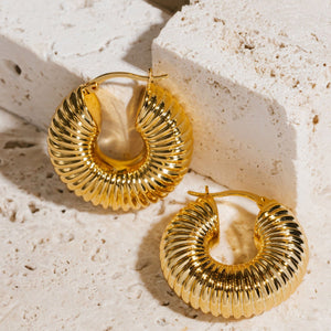 Two Brioche Hoop earrings lay side-by-side on a counter, the golden ridges shining where they catch the light.