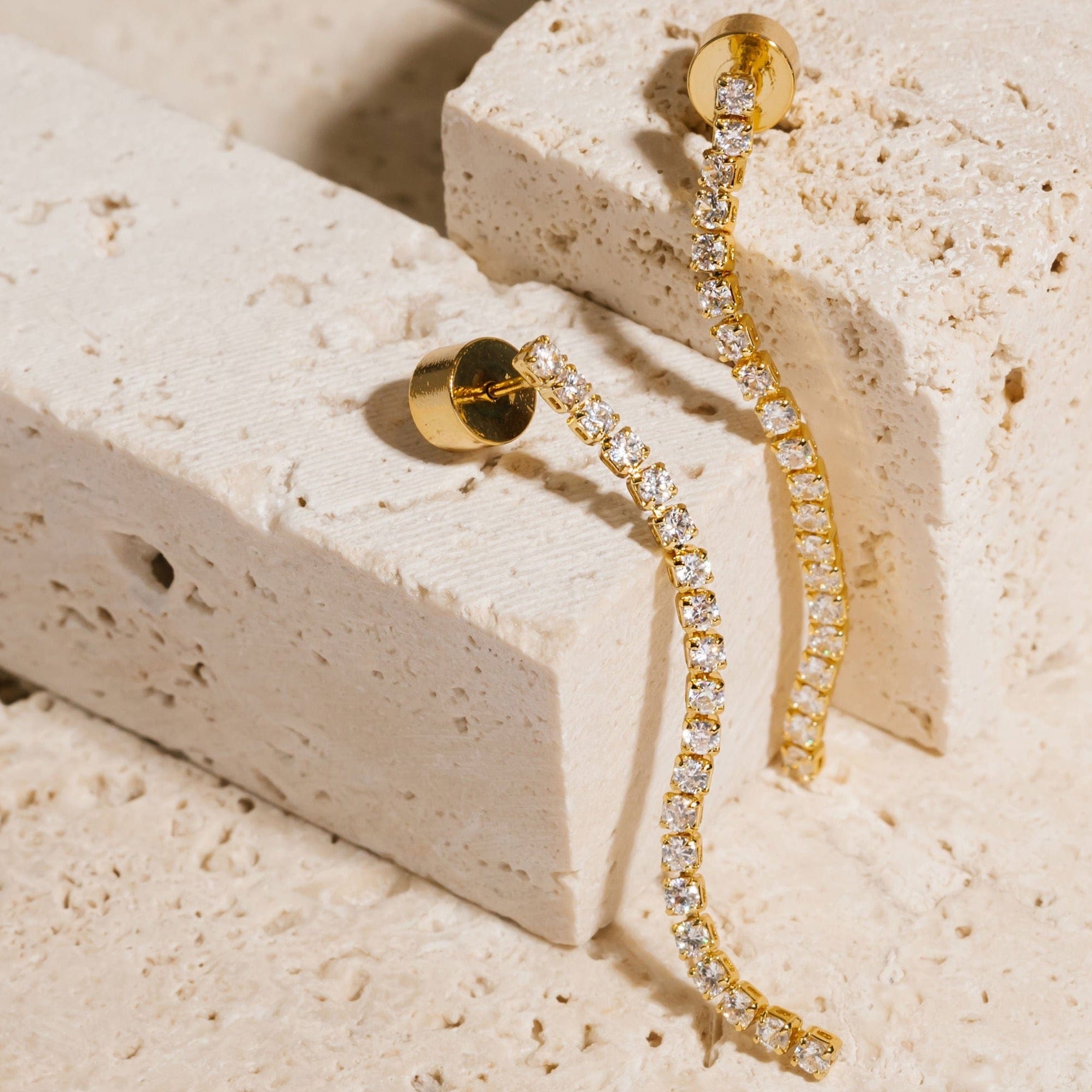 A pair of Scintilla Dangle Hoop earrings are set next to each other atop porous stone blocks, the cylindrical push-back closure shining golden behind the string of cubic zirconia accents. 
