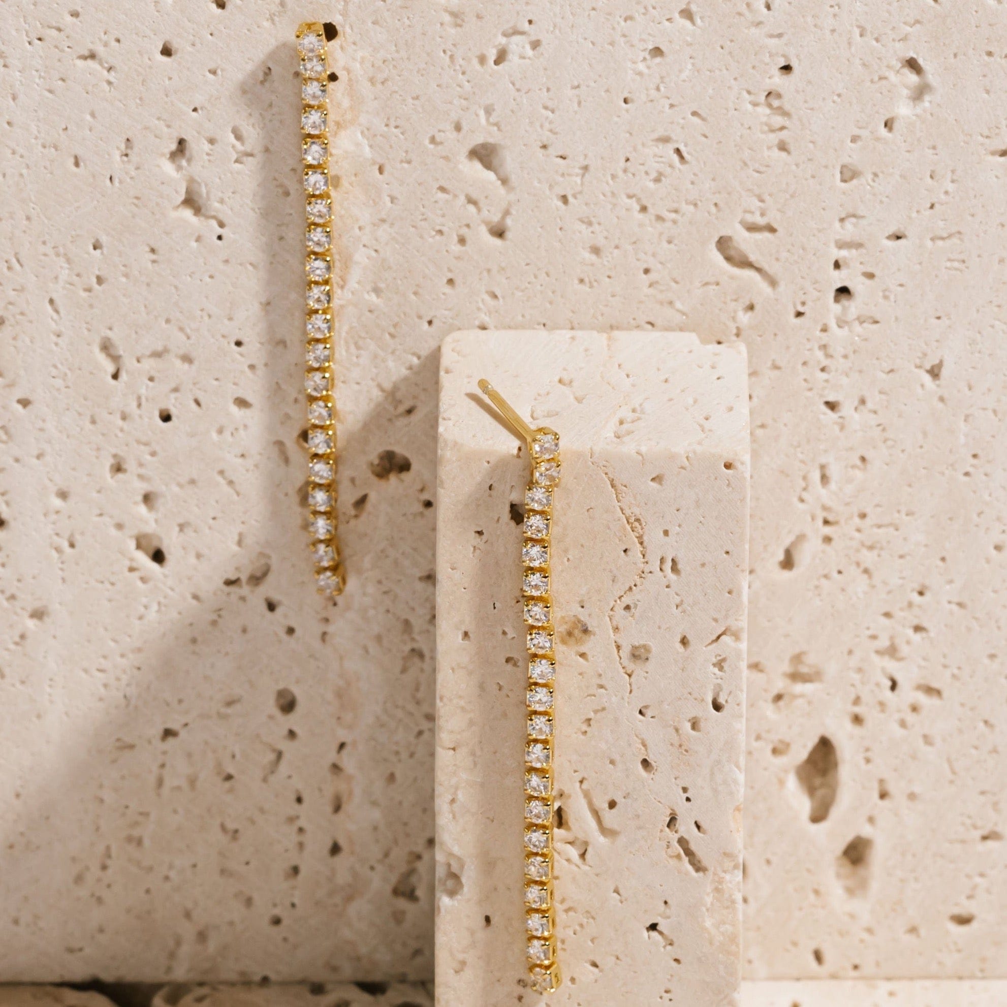 A Scintilla Dangle Hoop earring hangs from the side of a porous stone slab while a second earring cascades down a smaller stone block. 