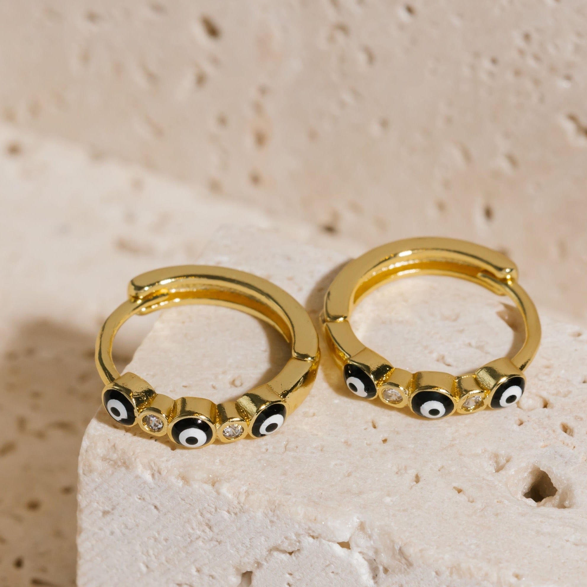 A pair of Evil Eye Hoop Noir earrings lay next to each other on a stone slab, the black and white evil eye symbols standing out against the golden hoop and subtle crystal accents. 