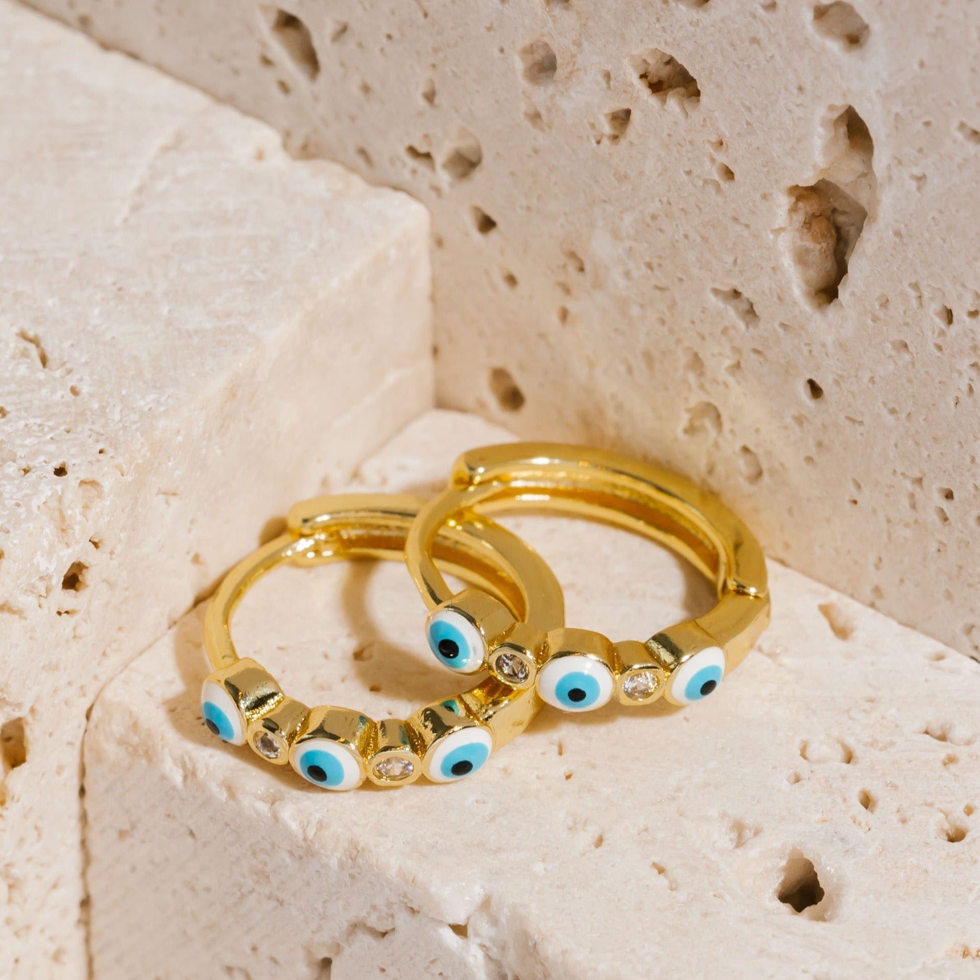A pair of teal Evil Eye Hoop earrings lay on stone blocks, one earring propped on the other as the evil eye symbols and crystal accents stare outward from the golden hoop