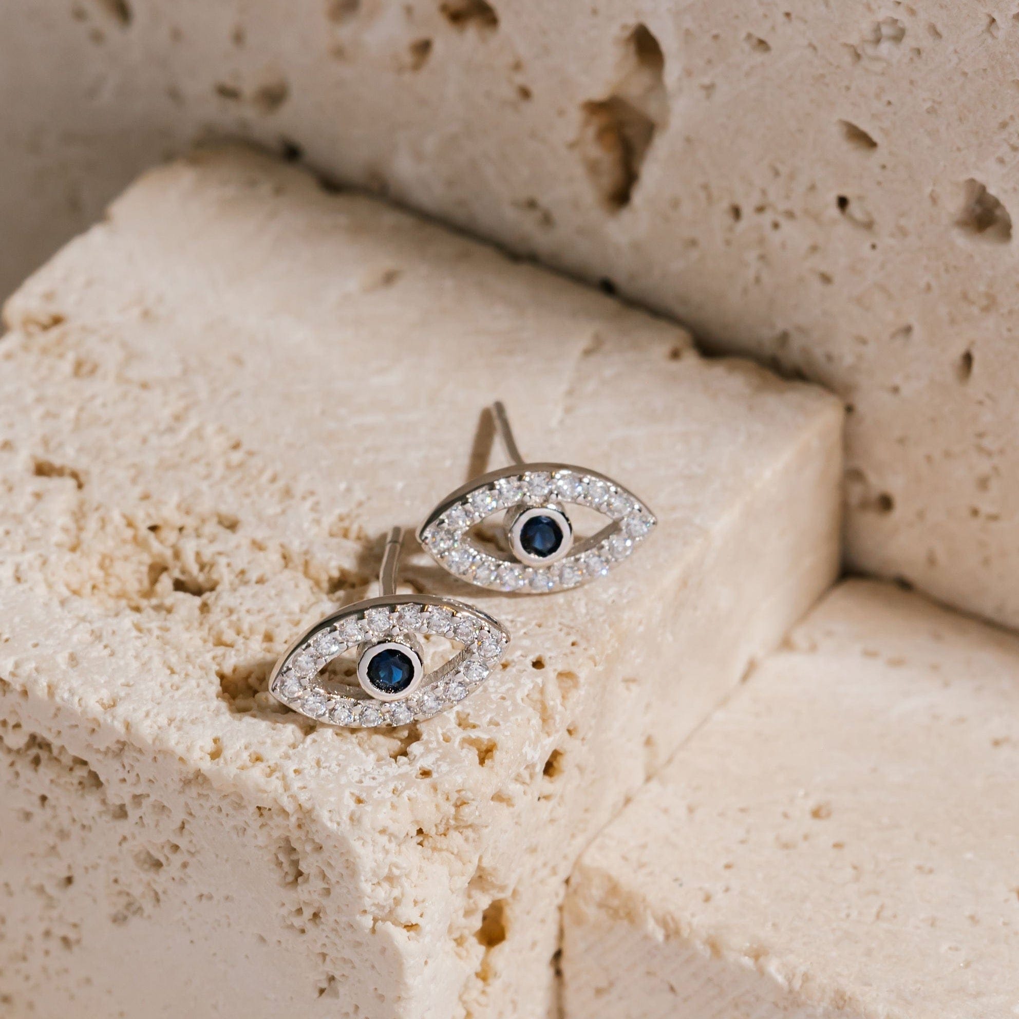 A pair of sterling silver Evil Eye Cristallo Stud earrings lay side-by-side on a  porous stone block, the blue sapphire and clear crystals sparkling in the light. 
