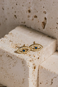 Two Evil Eye Cristallo Stud earrings lay next to each other on a beige stone block, the blue sapphire and crystal accents sparkling brightly against the golden eye-shaped earring. 