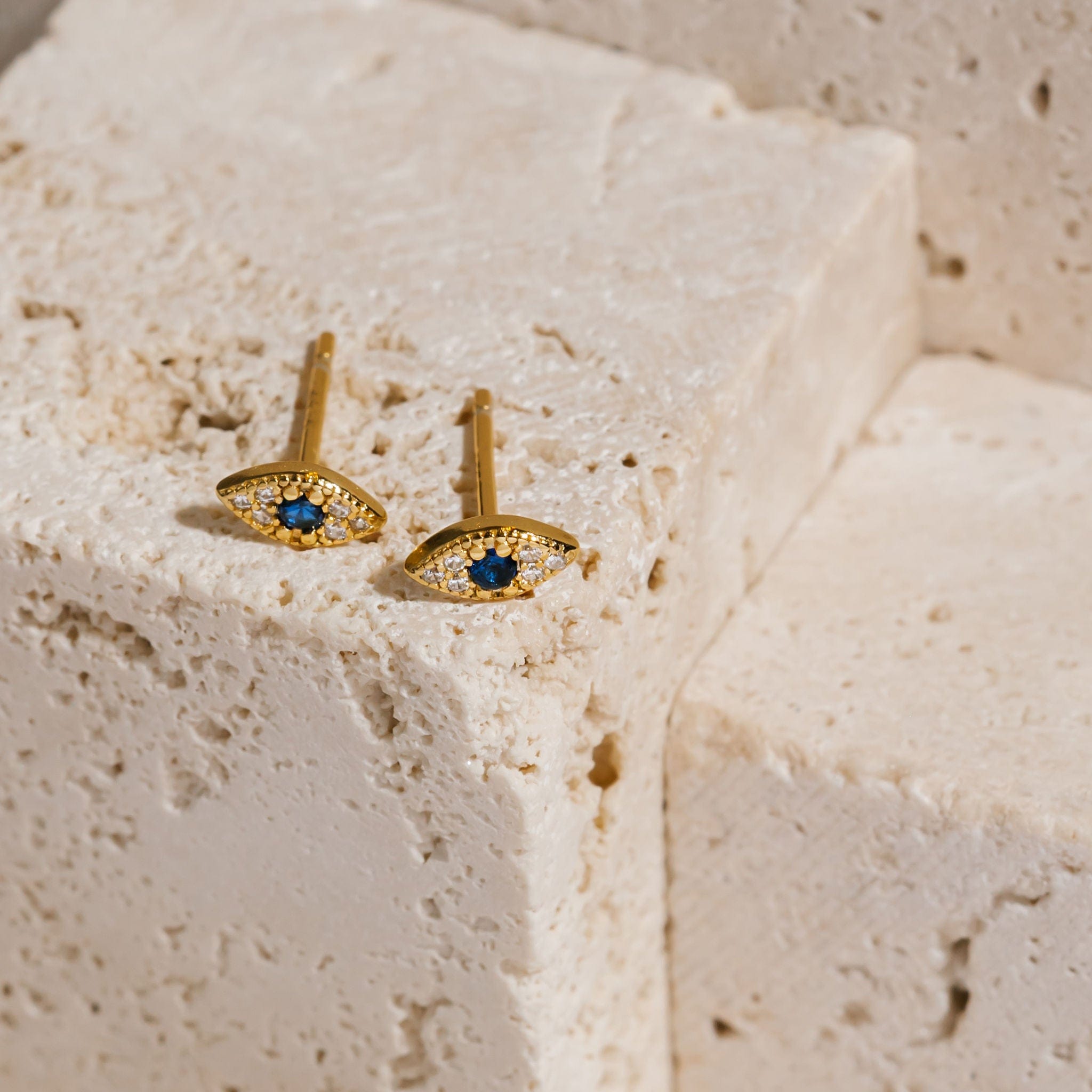 A pair of mini Evil Eye Cristallo Stud earrings lay side by side on a porous stone block, the golden texture of the earring highlighted amid the crystal and sapphire accents. 