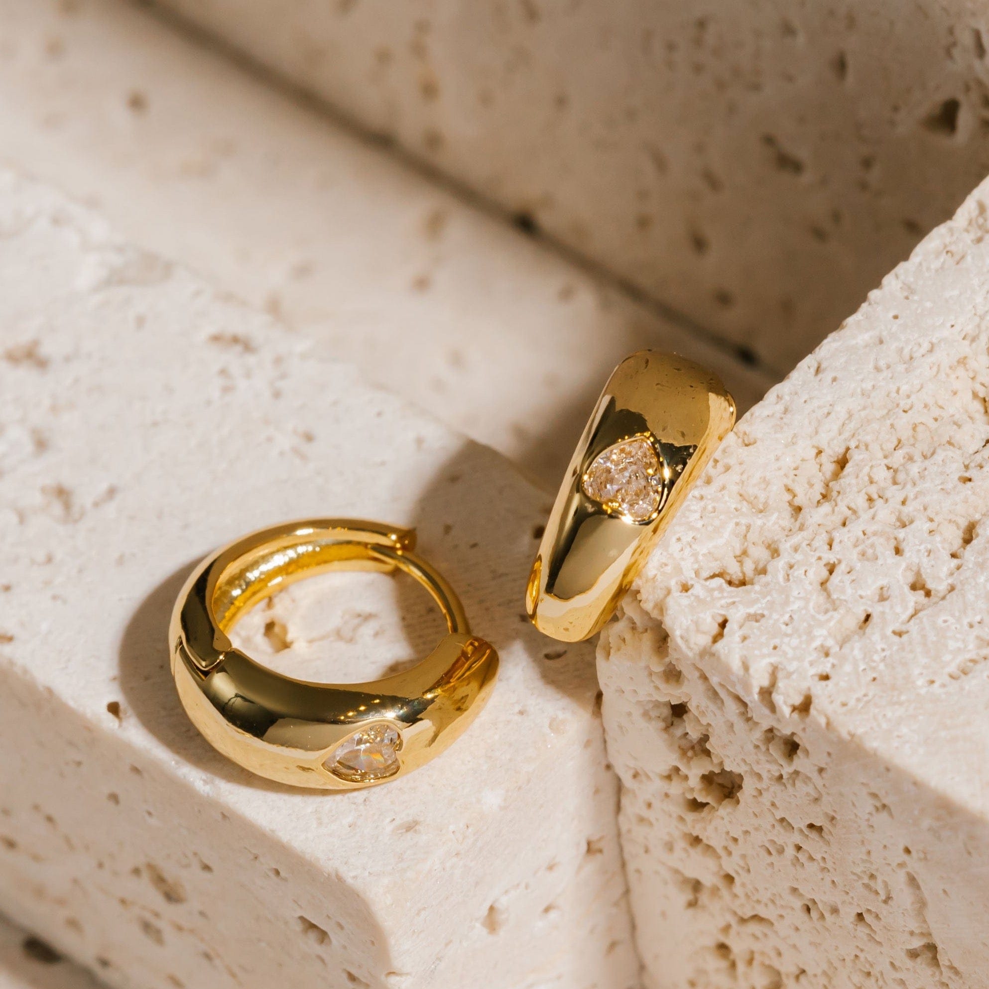 A pair of Amore Capri Hoop Mini earrings lay next to each other, one flat on its side and the other propped up to show off the sparkling heart-shaped crystal .  