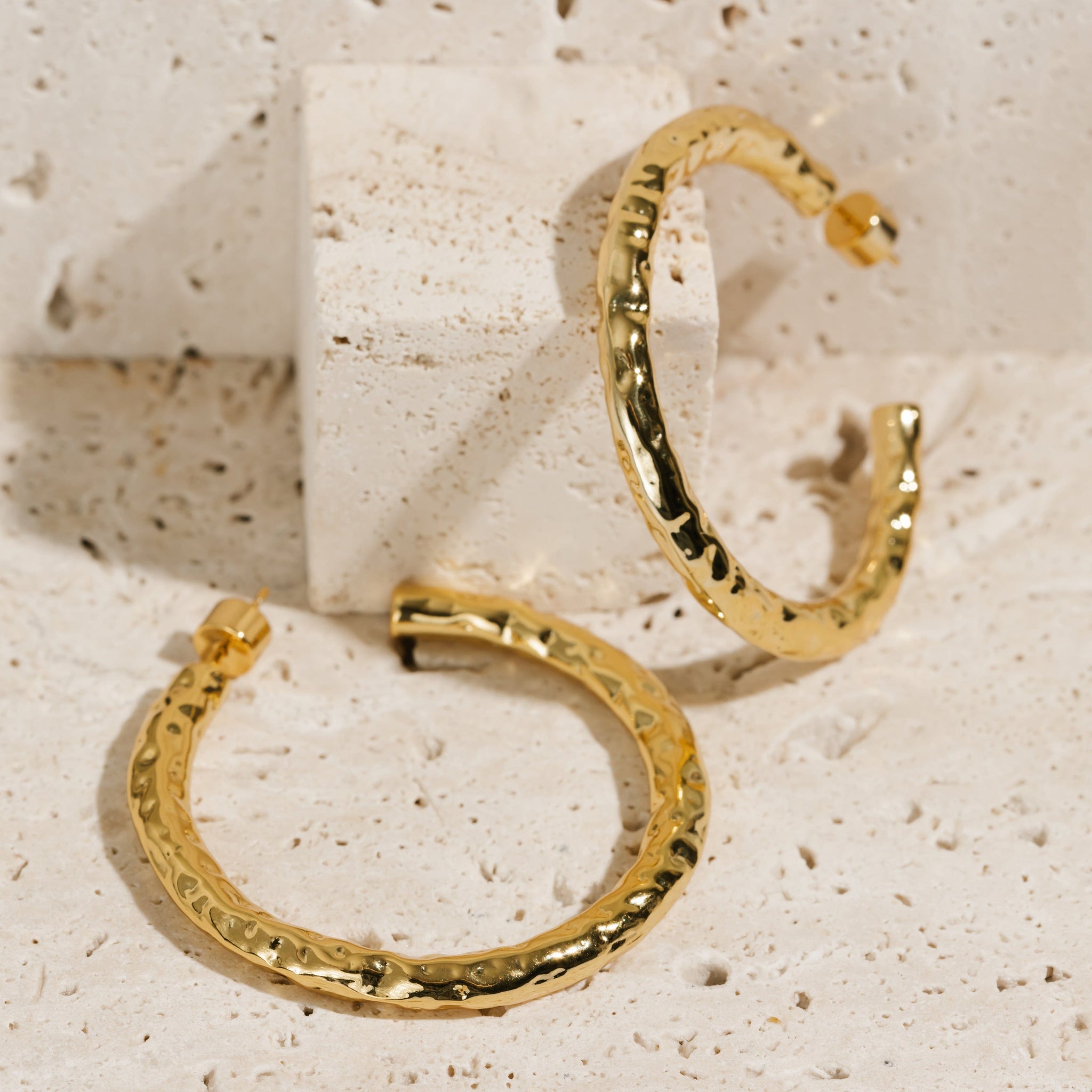 A pair of Marbella Hoop earrings is displayed on a stone slab, one earring laying on its side while the other is propped up. The various shades created by the hammered texture can be seen as the display light creates reflections and shadows on the open hoop earrings.
