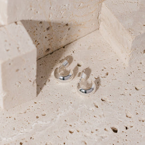 A pair of sterling silver Nice Hoop earrings sits on a porous stone surface, showing off the cylindrical push-back closure and sleek, open hoop design.