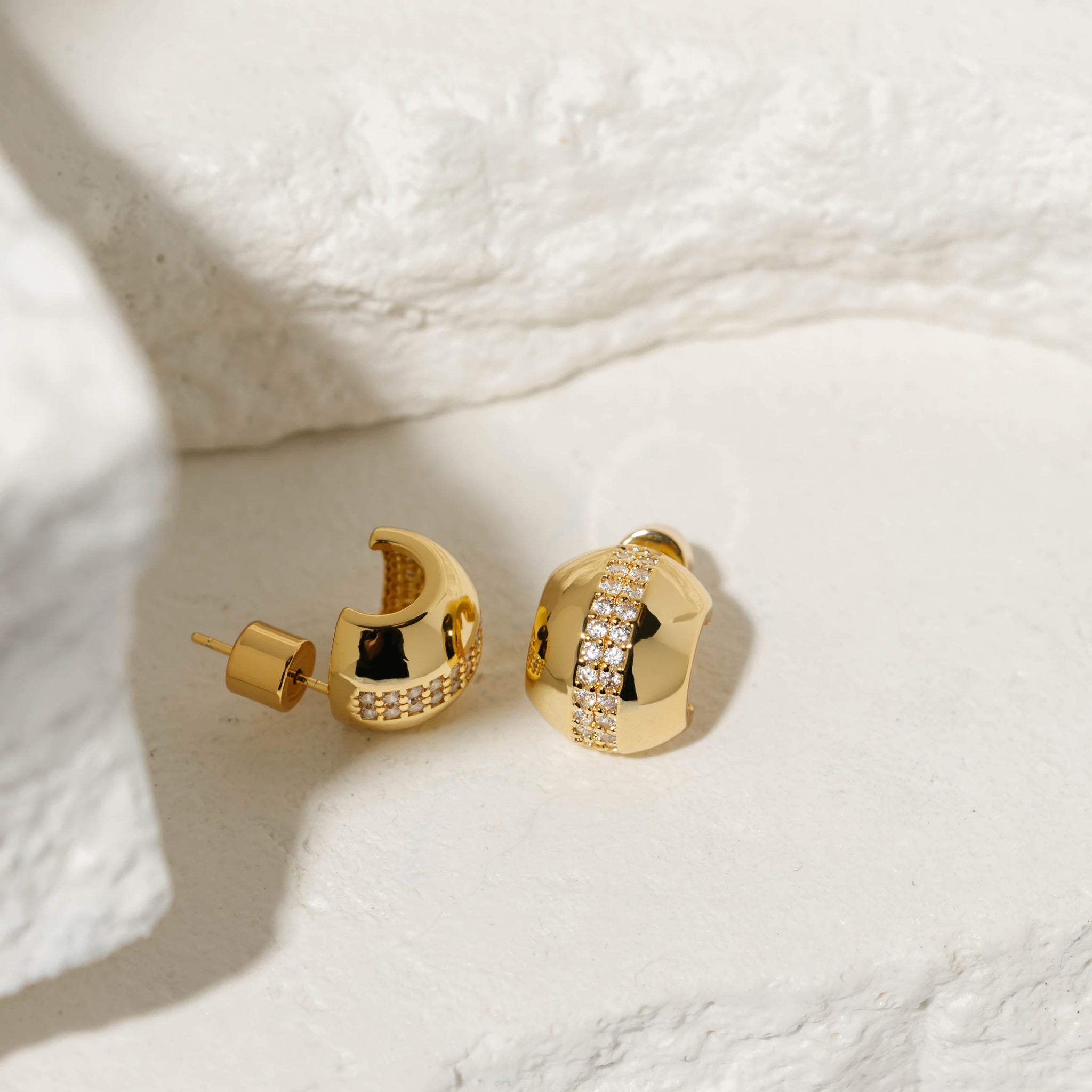 A pair of Nice Hoop Scintilla earrings sits on a stone slab, the rows of crystal accents sparkling upon the smooth, golden surface. 