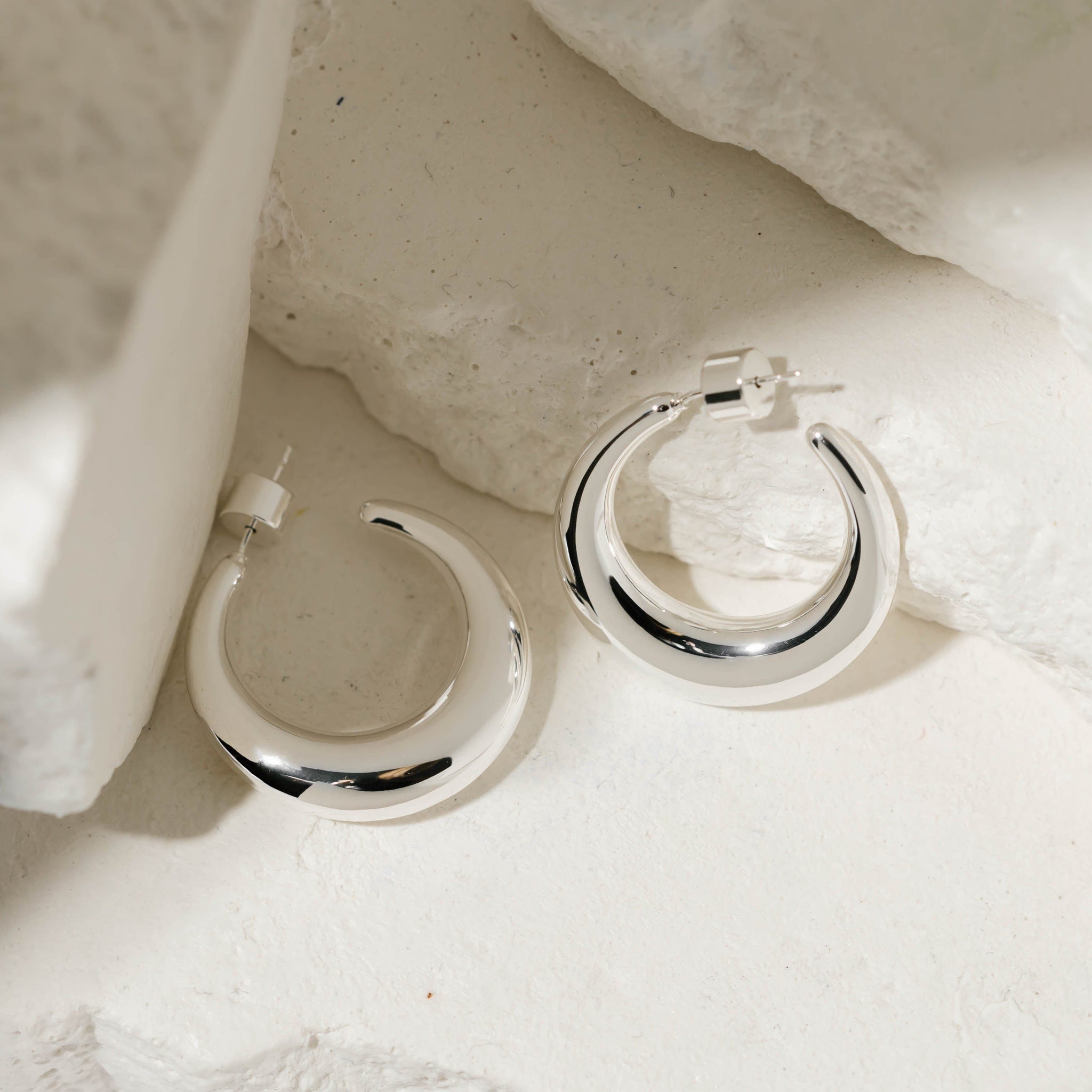 The crescent shape and cylindrical push back closures of a pair of sterling silver Mezzaluna Hoops are clearly displayed as the earrings lay against some white stone slabs. 