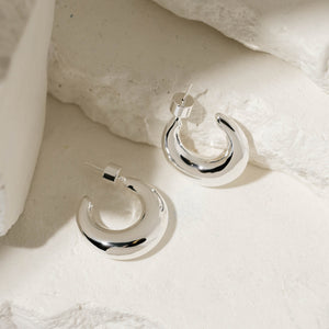 One sterling silver Mezzaluna Hoop Midi lies flat on its side while its pair is propped up against a white stone slab, both earrings displaying their crescent shapes and cylindrical push back closures. 