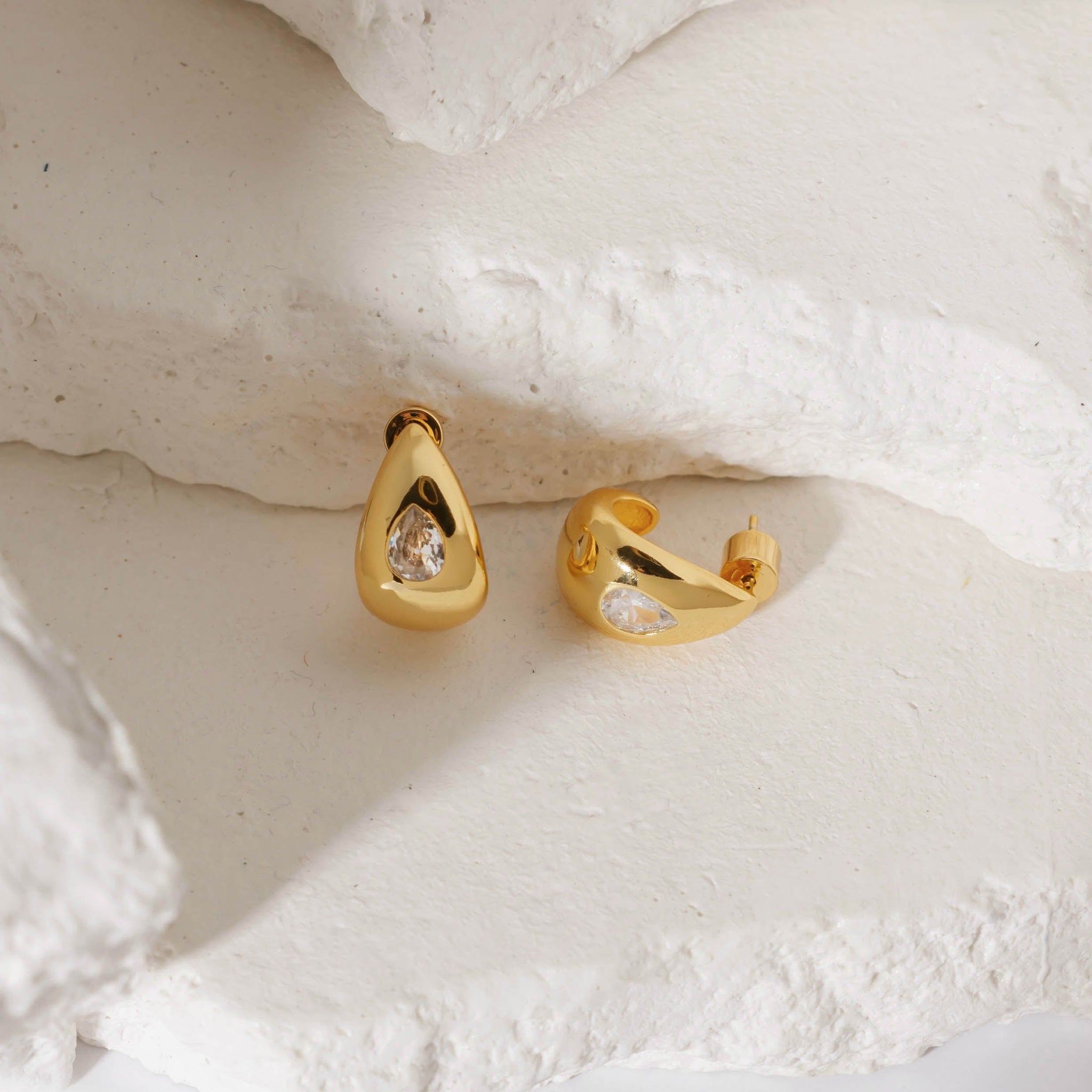 A pair of Amadora Hoop earrings with crystal accents on white stone slabs, one earring standing tall on its curve as the other lay on its side. 