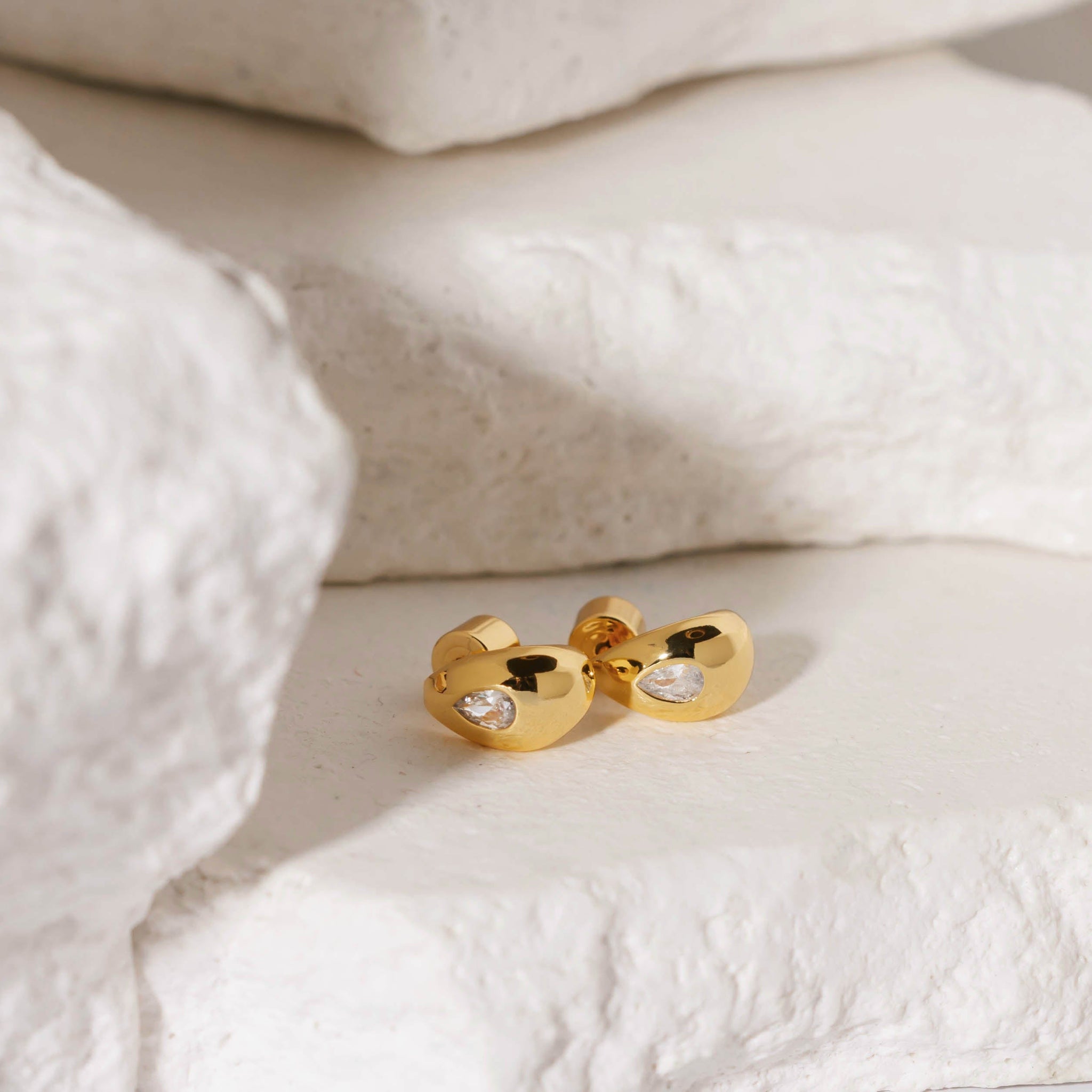 Two Amadora Huggie earrings with crystals lay together on a stone slab, the teardrop-shaped accents sparkling as the golden curves reflect their surroundings. 