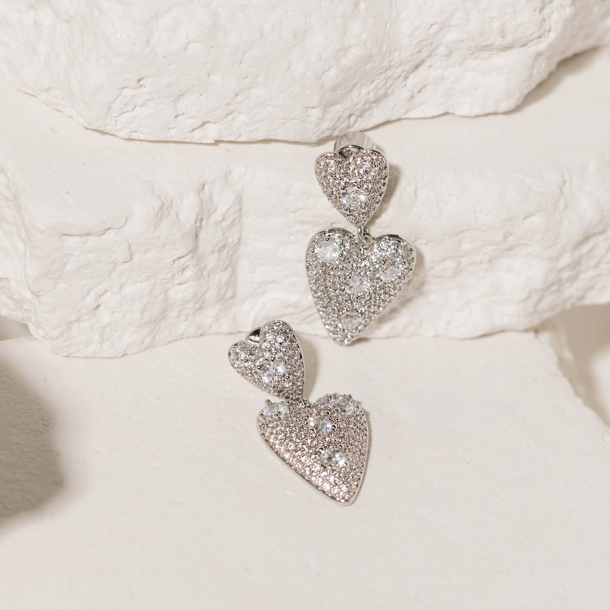 A pair of heart-shaped and crystal-adorned Cupid Drop Hoops sparkles brightly as the earrings are displayed against white stone slabs. 