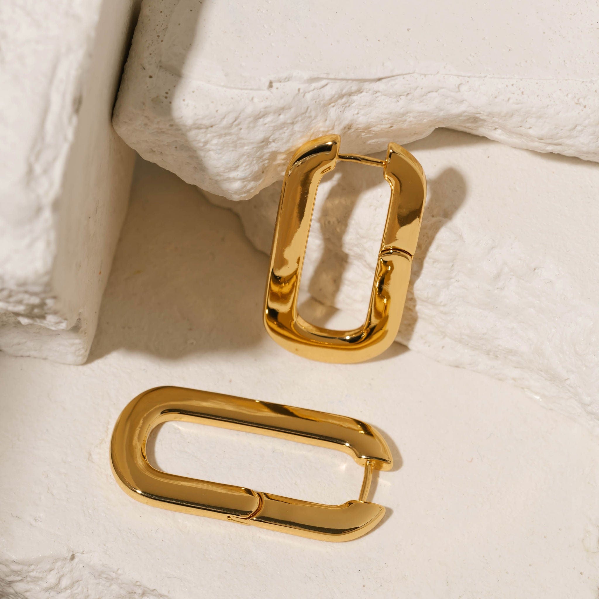 Positano Hoop Grande earrings in gold with an elongated rectangular shape and smooth, polished surface, elegantly arranged on a textured stone background. These statement hoops by The Perfect Hoop offer a chic, contemporary style