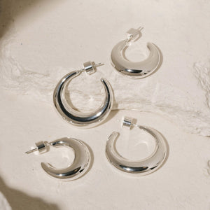 The two pairs of earrings in the Mezzaluna Hoop Duo sit atop white stone slabs, showing a size comparison of the two crescent shaped hoop earrings. 