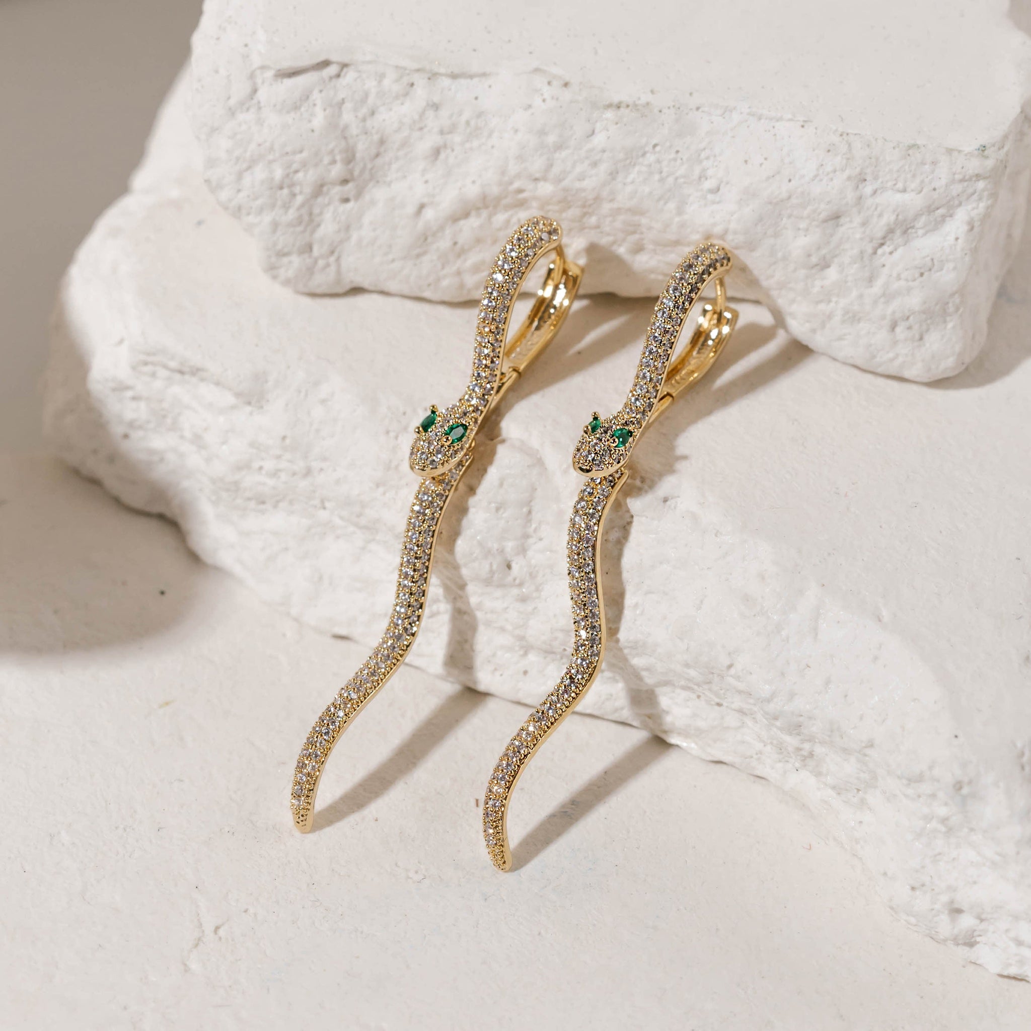 A pair of long, serpentine Eden Hoop earrings sits against tiered stone slabs, the crystal-covered bodies sparkling brightly.