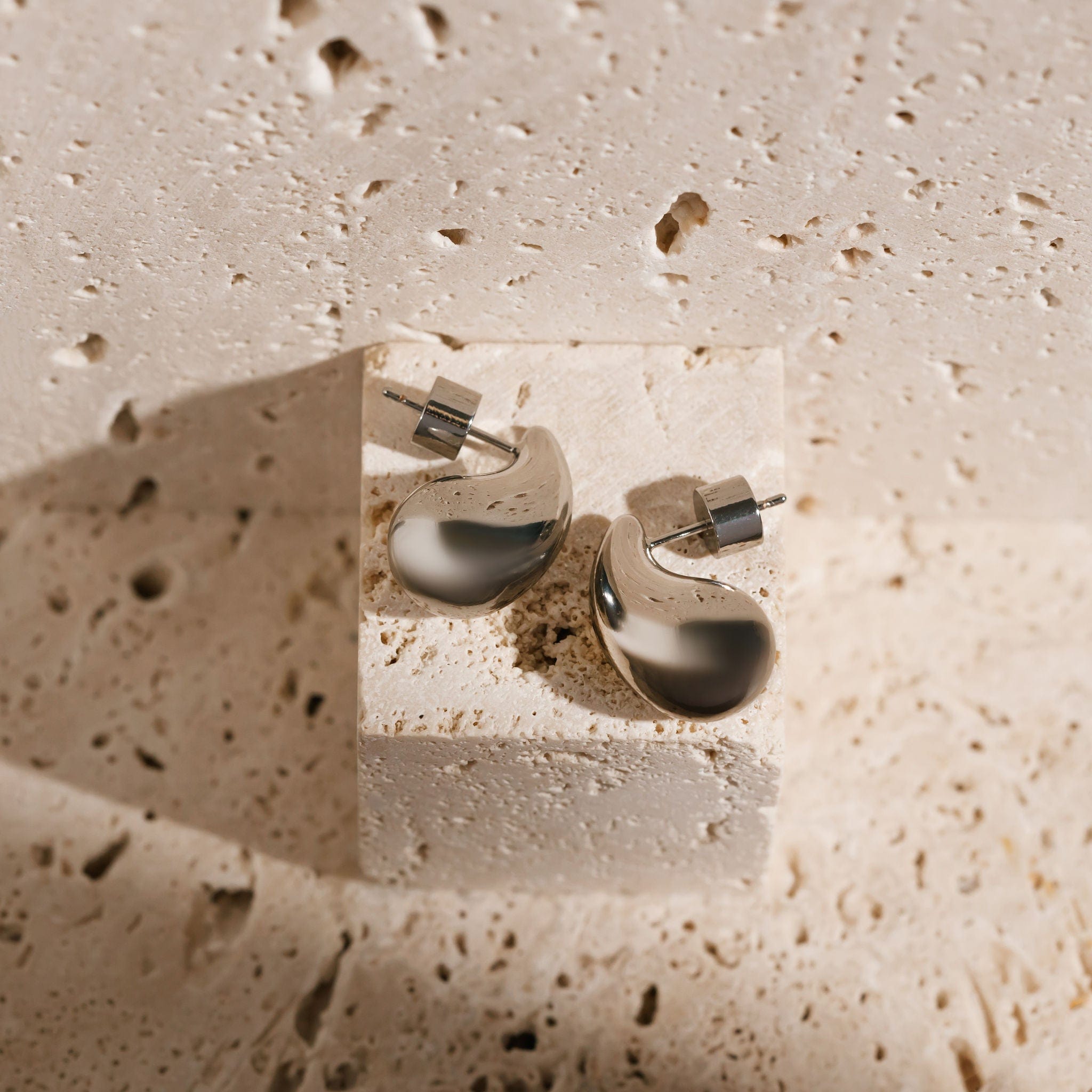 A pair of platinum Moda Hoop Mini earrings is displayed on a porous stone block, showing off the teardrop-shaped open hoop design and the cylindrical push-back closures. 