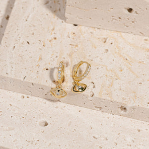 Two Evil Eye Solitaire Drop Hoop earrings sit side by side on a stone slab, the golden drop hoop earrings glimmering as the crystal accents atop the hoops and in the center of each evil eye charm sparkle brightly. 