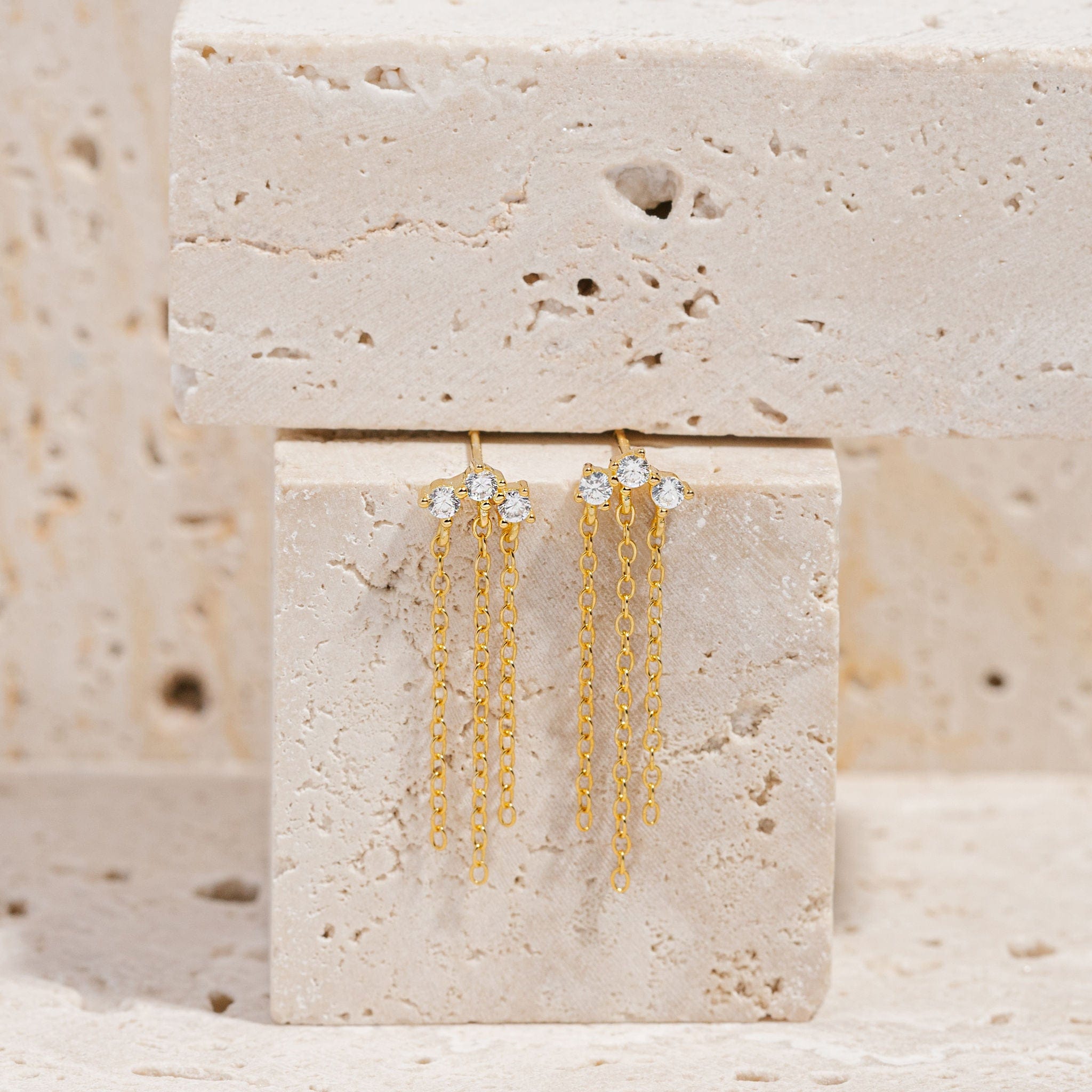 A pair of Stella Chain Drop Stud earrings lay suspended on a stone block, the delicate golden chains cascading gracefully down from each of the crystal accents. 
