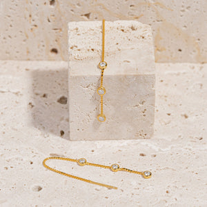 A pair of Coquille Chain Threader earrings is displayed,  one earring laying flat in a U-shape as the other hangs gracefully across a stone block. 