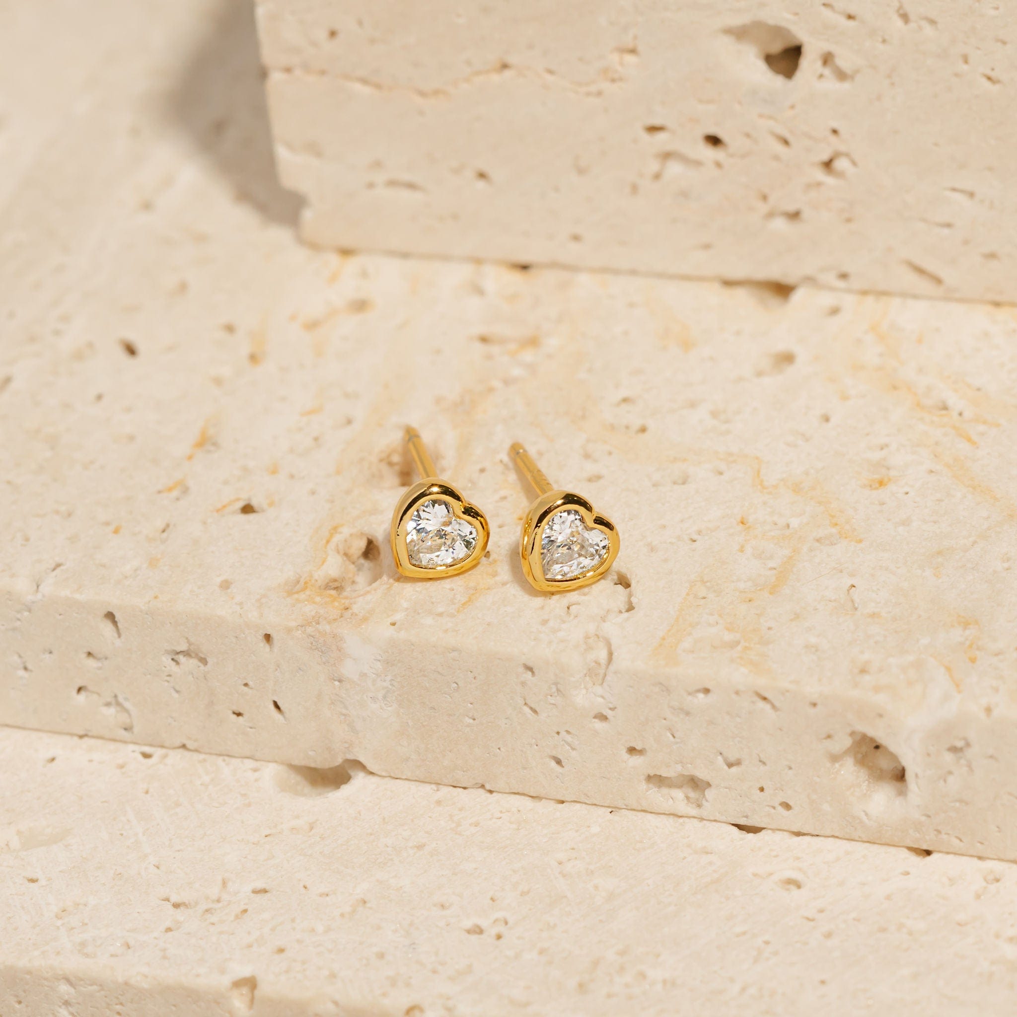 A pair of Amore Stud earrings lays side by side on a stone slab, the crystal accents sparkling bright within the heart-shaped settings. 