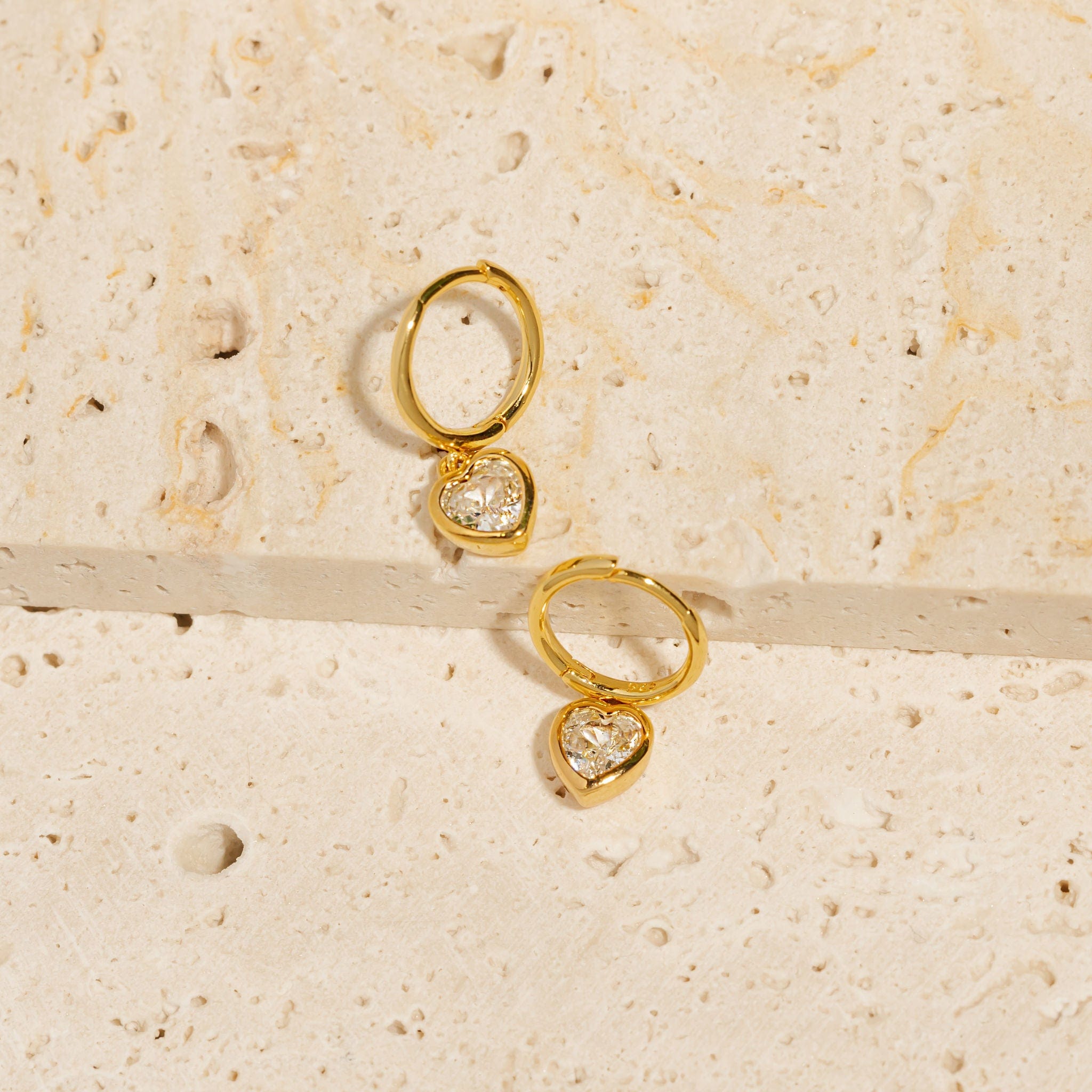 A pair of Amore Solitaire Drop Hoop earrings lay on porous stone slabs, the round golden hoops and heart-shaped crystal charms gleaming where they catch the light. 