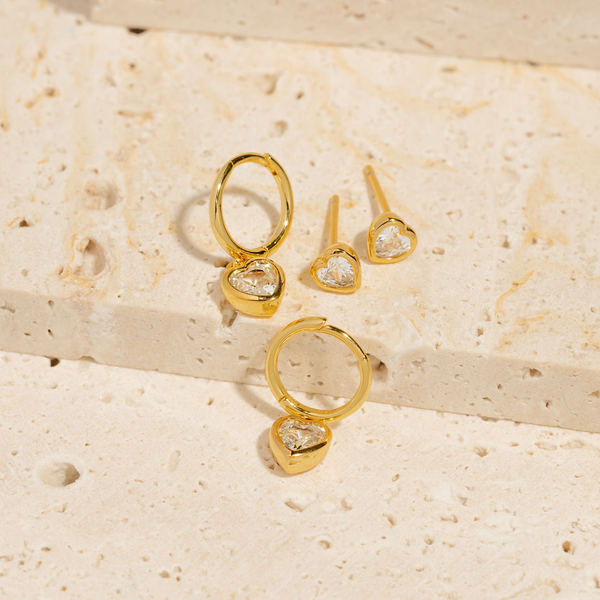 A pair of Amore Drop Hoop earrings is displayed next to a pair of Amore Stud earrings, each showing off its heart-shaped crystal accents and shining gold settings. 