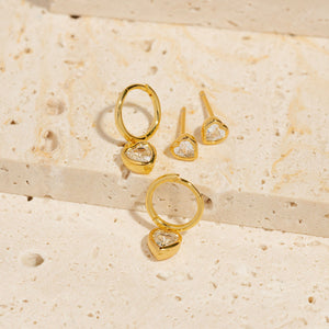 A pair of Amore Drop Hoop earrings is displayed next to a pair of Amore Stud earrings, each showing off its heart-shaped crystal accents and shining gold settings. 