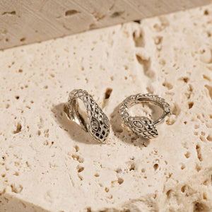The snake-like design, crystal eyes, and lever back closure of a pair of Veleno Huggie earrings can be seen from two angles as one earring is propped up and the other lays on its side.  