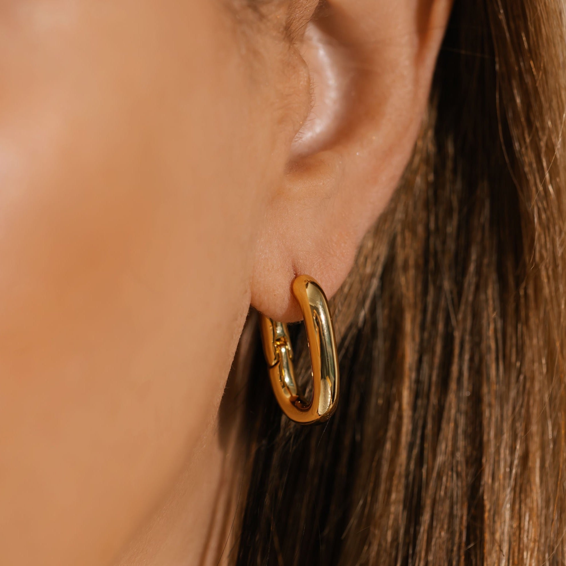 A close-up of The Positano Hoop earring worn on the ear, highlighting its bold, elongated oval form and glossy gold surface.  By The Perfect Hoop.
