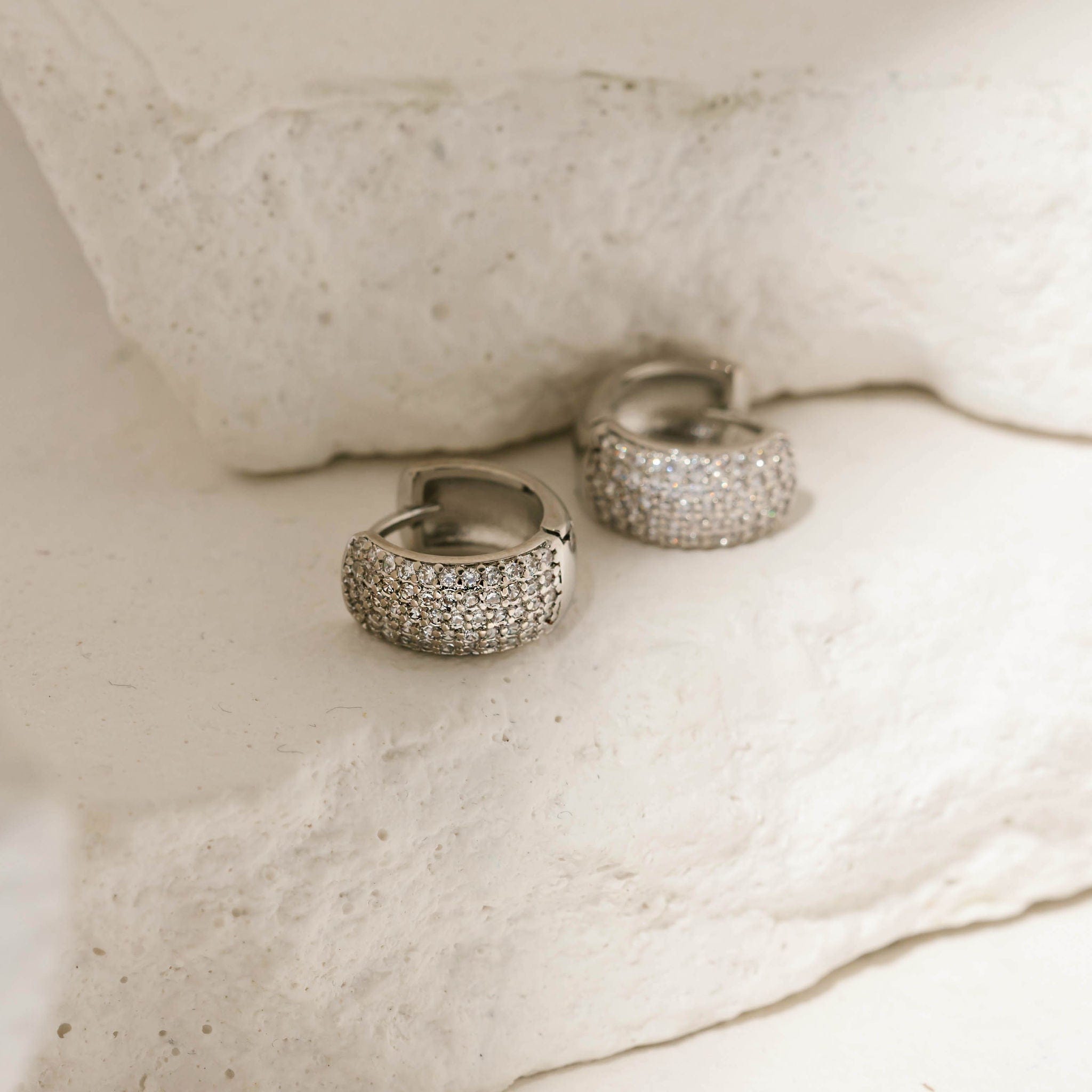 White gold pavé Sicily hoop earrings with a wide band encrusted with sparkling crystals, elegantly displayed on a textured stone surface. These luxurious pavé hoop earrings by The Perfect Hoop offer a bold and refined look.