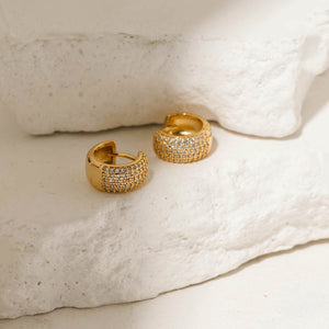 Gold pavé Sicily hoop earrings with a wide band encrusted with sparkling crystals, displayed on a textured stone surface. Elegant and bold, these hoops by The Perfect Hoop add a statement look.