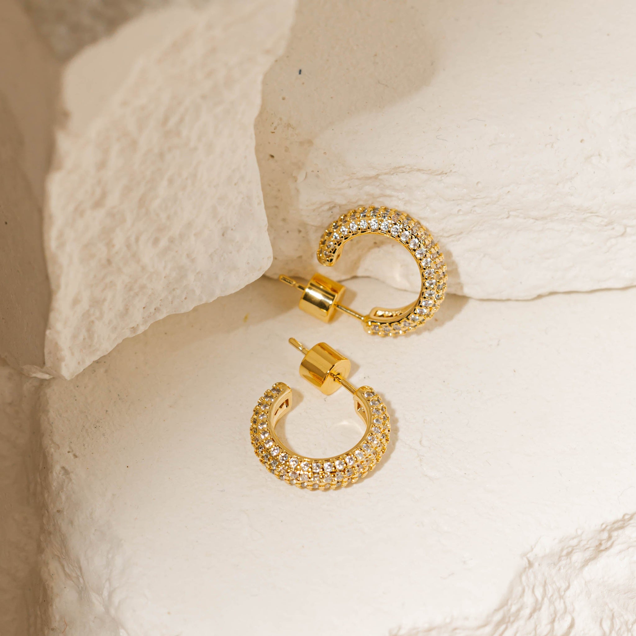 The open hoop design, cylindrical closures, and crystal accents of a pair of Siena Hoop Mini Pavé earrings are displayed against white stone slabs.