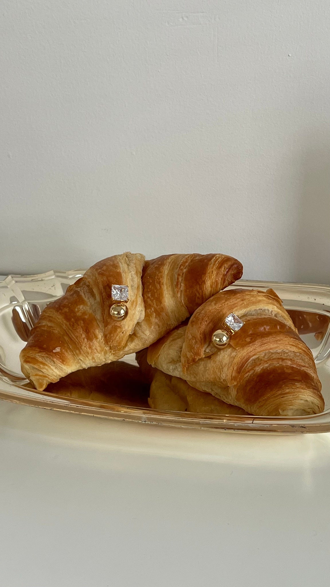 Two croissant-shaped pastries with The crystal and gold Sofi Stud by The Perfect Hoop on a silver tray and  white surface.