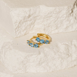 A pair of Something Blue Cristaux Huggie earrings is displayed, one earring sitting atop the other while their gold hoops and blue crystal accents shine in the light. 