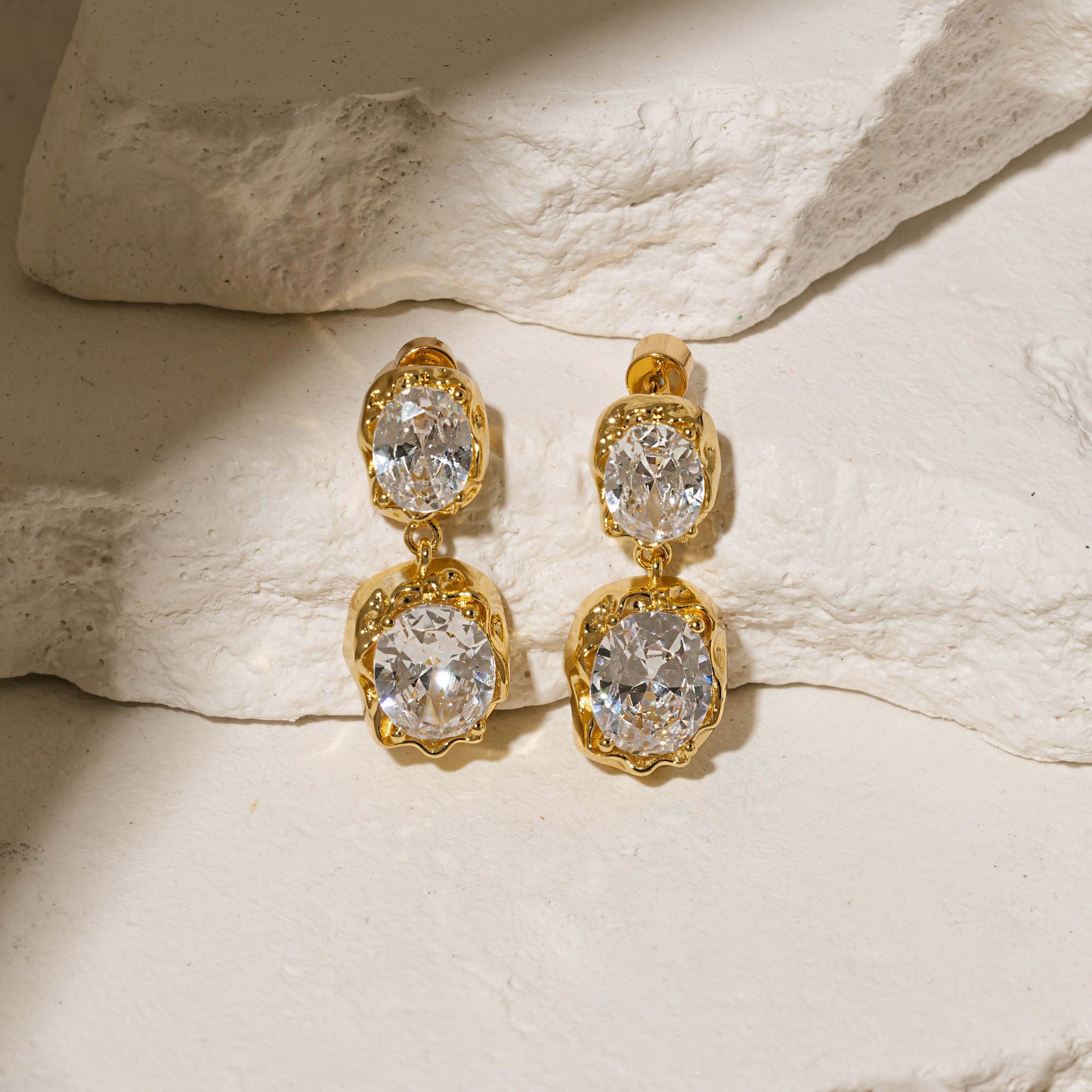 The oval crystals and gold settings of a pair of Aura Casila Hoop earrings sparkle brightly under the spotlight as the earrings sit propped against white stone slabs. 