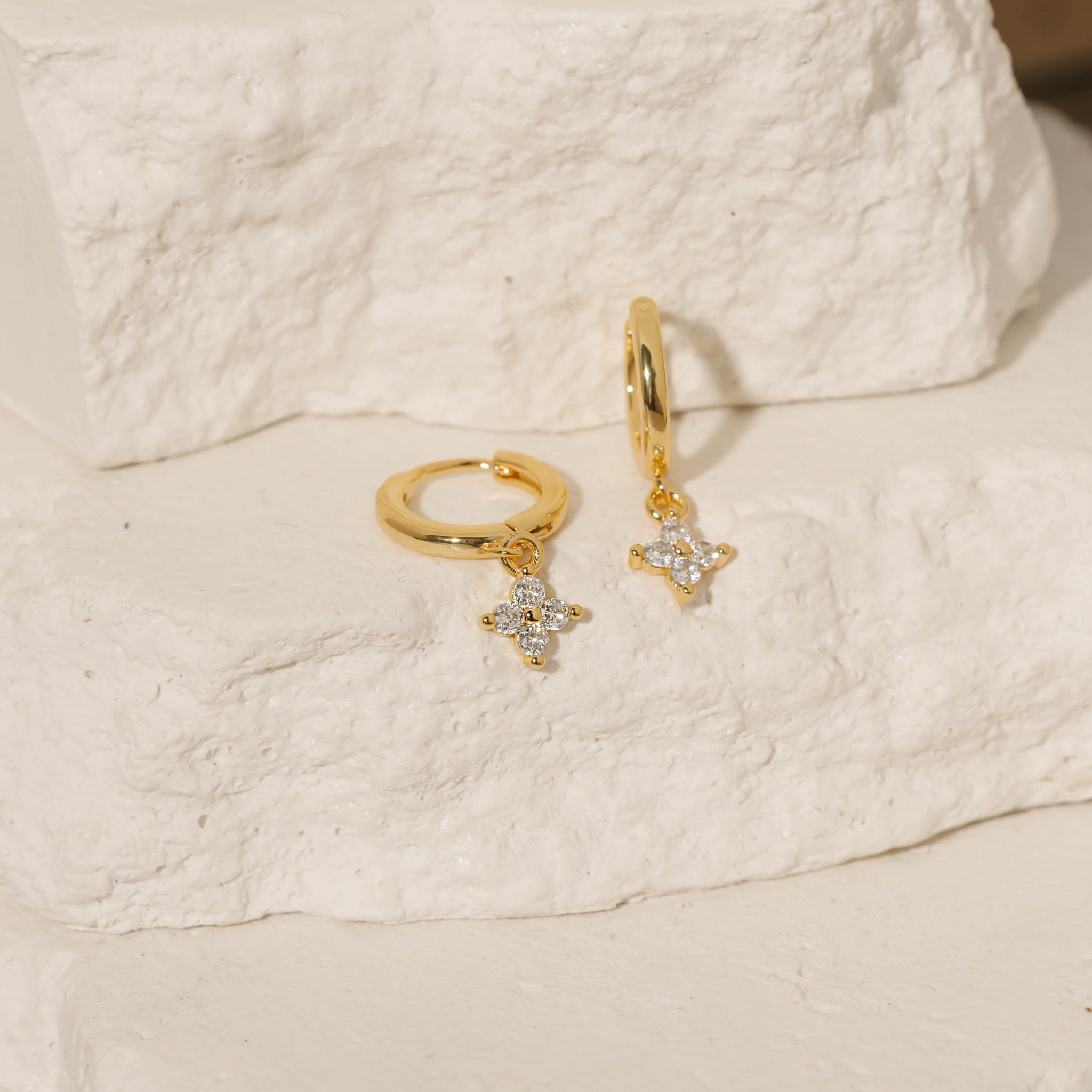 Two Clover Drop Hoop-Crystal earrings are displayed on tiered stone slabs, each gleaming from its sleek golden hoop and clear crystal accents. 