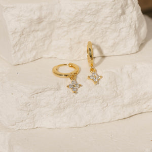 Two Clover Drop Hoop-Crystal earrings are displayed on tiered stone slabs, each gleaming from its sleek golden hoop and clear crystal accents. 